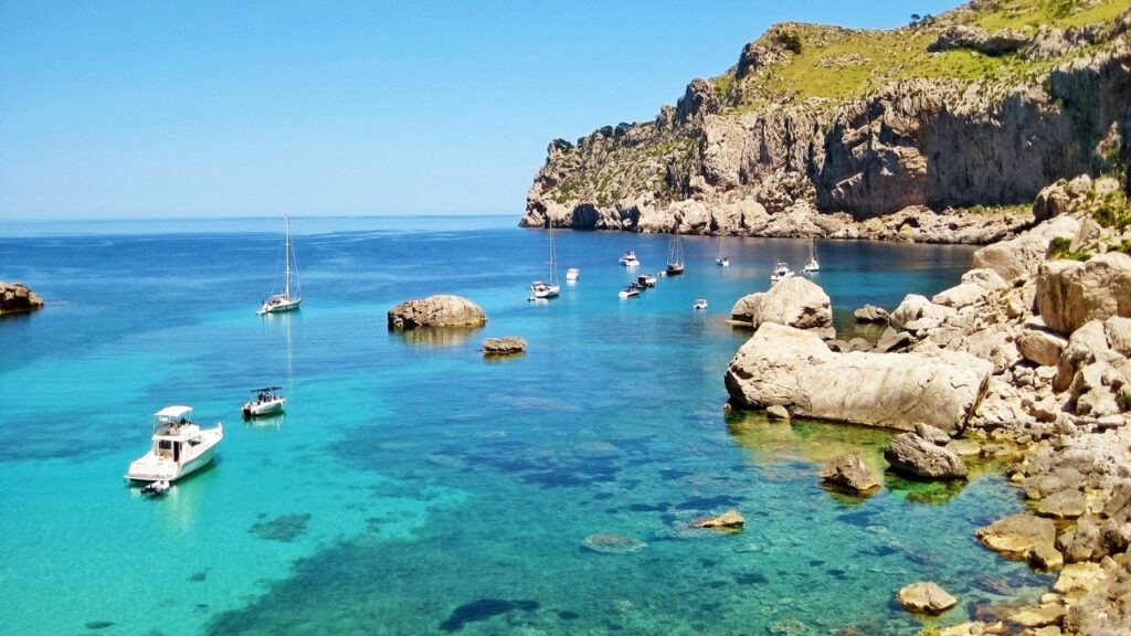Panoramic view of Mallorca’s coastline with turquoise waters, sandy beaches, and Mediterranean buildings surrounded by green hills.